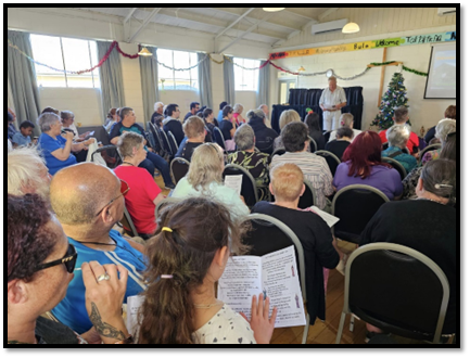 A large group of seated people singing Christmas Carols.