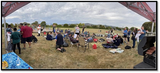 A large group of people sitting on the grass at a reserve beside a playground.
