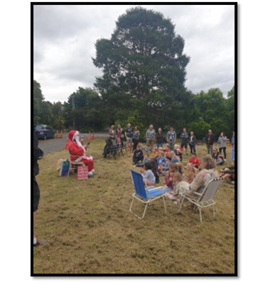 Santa Claus seated outside in front of a group of people seated and standing on a grassed area.
