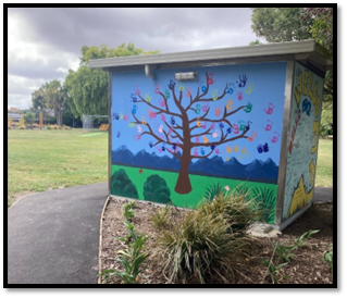 A mural of tree painted in bright colours with the leaves as coloured hand prints on the side of a small toilet block.


