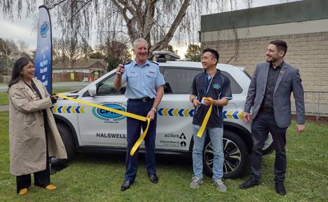Four people standing outside in front of the new patrol car with Senior Sergeant Roy Appley, cutting a yellow ribbon.
