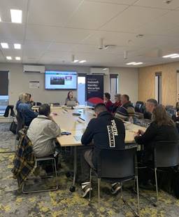 A group of people sitting around tables listening to a presentation, displayed on a screen at the back of the room.

