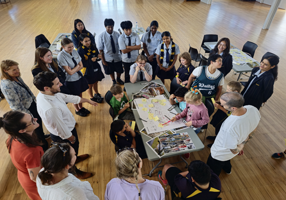 A group of students and adults standing around  table having a discussion/participating in a workshop