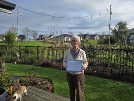 Karen Whitla standing outside, holding her certificate, with green space and houses in the background