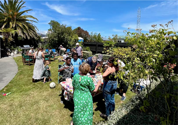 Photograph of a backyard gathering with Oaklands residents socialising around a table and others sitting on outdoor furniture. The scene includes a large palm tree, and a clear blue sky, indicating a sunny day event.
