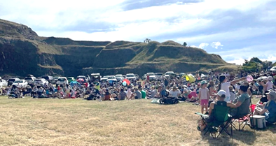 Photograph at Halswell Quarry with people sitting on grass and folding chairs, surrounded by parked vehicles and hills in the background. Bright daylight and partly cloudy sky create a relaxed atmosphere.
