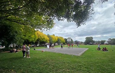 Photograph of an outdoor basketball court at Harrington Park, with several people playing and others watching or sitting on the grass. The court is surrounded with trees and the sky is overcast.