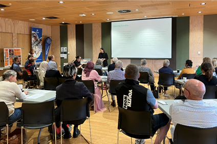 Photograph of the Hapori Hui in the Kahikatea Hall at Rārākau Riccarton Centre. Attendees are listening attentively, seated at tables facing a presenter and a large projection screen.