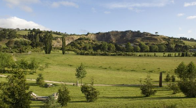 Photograph of Halswell Quarry Park, a large green area with scattered trees and a hill in the background under a partly cloudy sky.