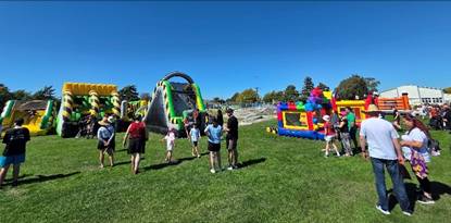 Photograph featuring inflatable slides and bounce houses on a grassy field under clear blue sky. Several people, including children and adults, are gathered around.