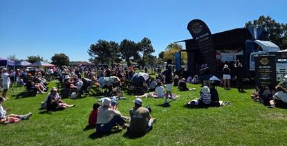 Photograph of a crowd sitting on grass facing a stage set up on a truck, under clear blue sky. The stage features black banners with gold text.