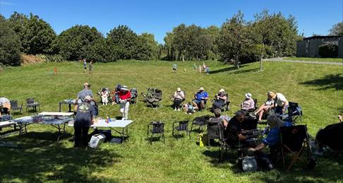 Photograph of an outdoor gathering in a grassy park area under clear blue sky, showing people sitting on folding chairs and standing around tables. 